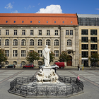Blick auf den Gendarmenmarkt in Berlin mit dem Schiller-Denkmal-Brunnen im Vordergrund und der Fassade der Berlin-Brandenburgischen Akademie der Wissenschaften im Hintergrund, daneben ein modernes Glas-Bürogebäude.