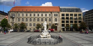 Blick auf den Gendarmenmarkt in Berlin mit dem Schiller-Denkmal-Brunnen im Vordergrund und der Fassade der Berlin-Brandenburgischen Akademie der Wissenschaften im Hintergrund, daneben ein modernes Glas-Bürogebäude.